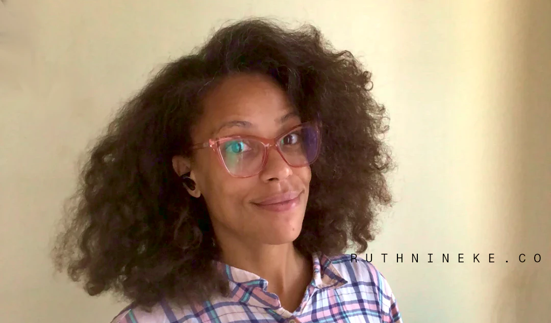A woman wears pink glasses and a pink plaid shirt as she stands before a wall smilling. Photo of Ruth Nineke, writer and web designer who provides small creatitve online businesses with SEO content strategy and more.